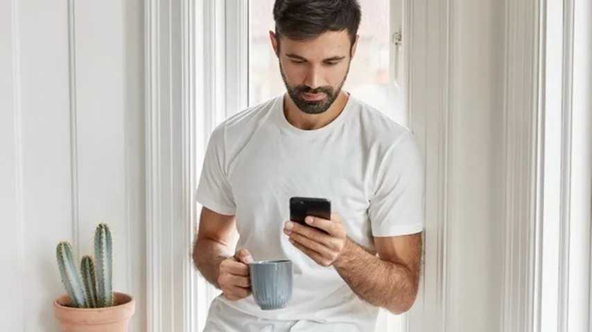 Hombre joven leyendo noticias con un smartphone mientras se bebe de una taza. (Foto: Freepik)